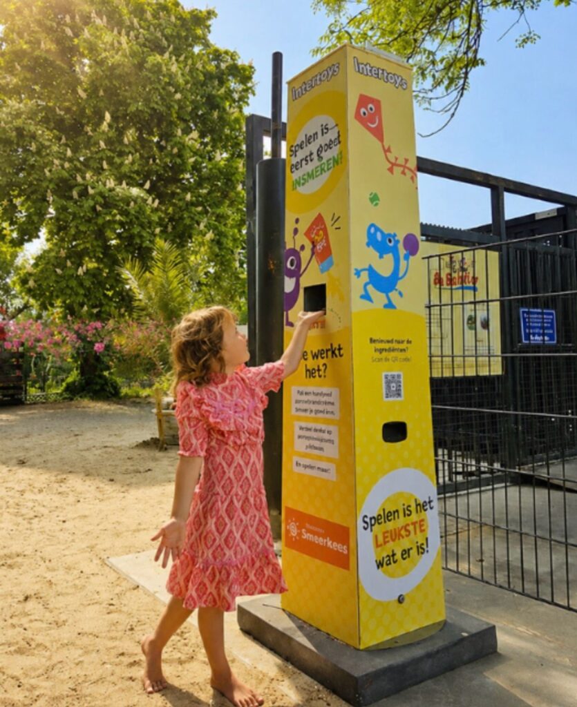 Mädchen holt sich kostenlose Sonnencreme an einer gelben Sonnencreme-Station im Intertoys-Design auf einem Spielplatz