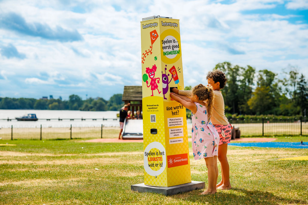 Two children are getting sunscreen at an Intertoys-themed sunscreen station on a playground by the lake