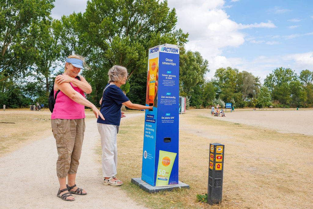 Two women are applying sunscreen at a sunscreen station set up by the non-food discount store Action in a park