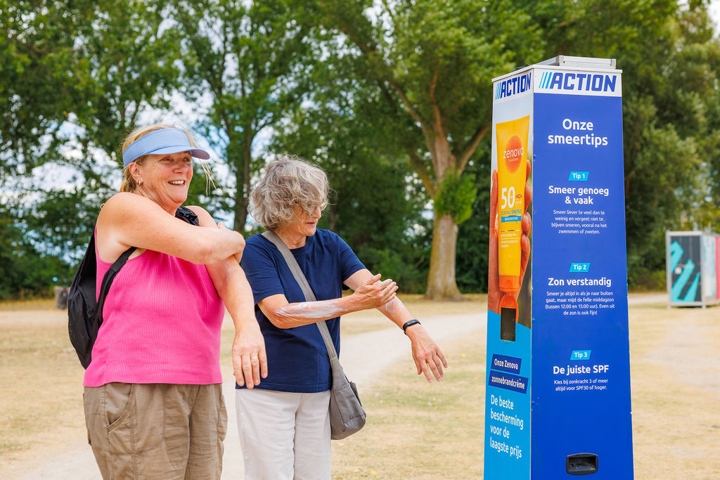 Zwei ältere Frauen cremen sich lachend die Arme mit kostenloser Sonnencreme ein, neben einer blauen Sonnencreme-Spendersäule von Action mit Zenova-Sonnencreme LSF 50 in einem sonnigen Park