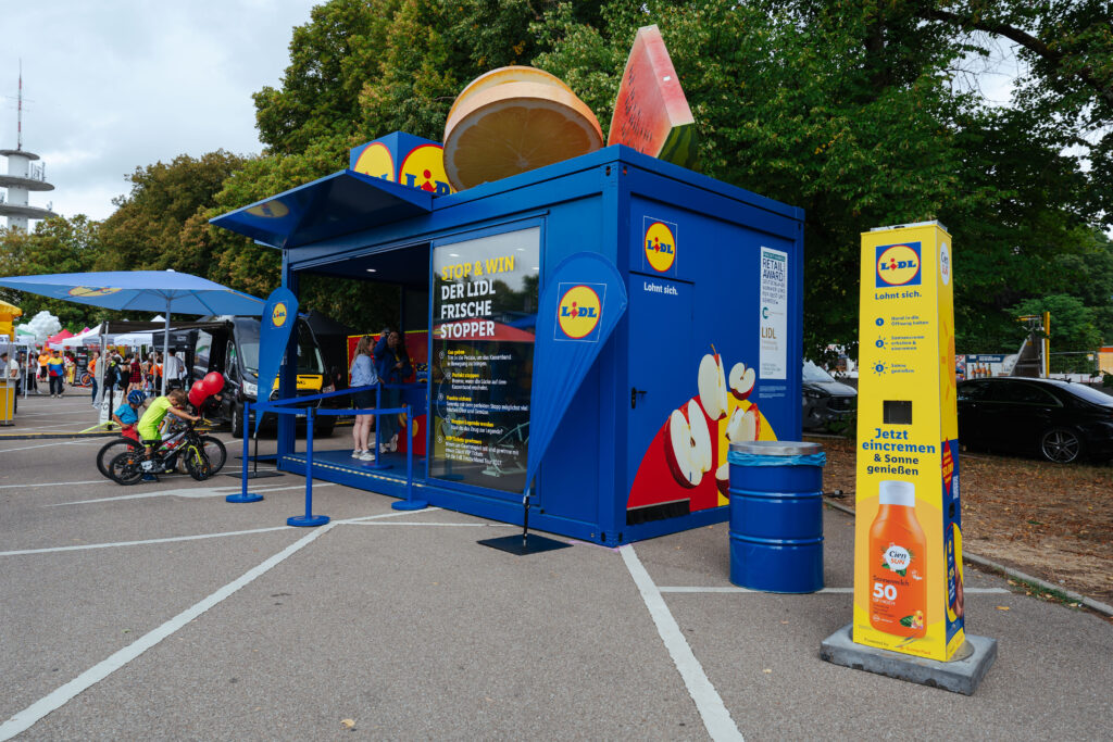 Gelbe Sonnencreme-Spendersäule von Lidl und Cien Sun, „Powered by Sunny Paul", neben einem blauen Lidl-Aktionsstand „Stop & Win – Der Lidl Frische Stopper" bei der Lidl Deutschland Tour, im Hintergrund Besucher und Kinder mit Fahrrädern