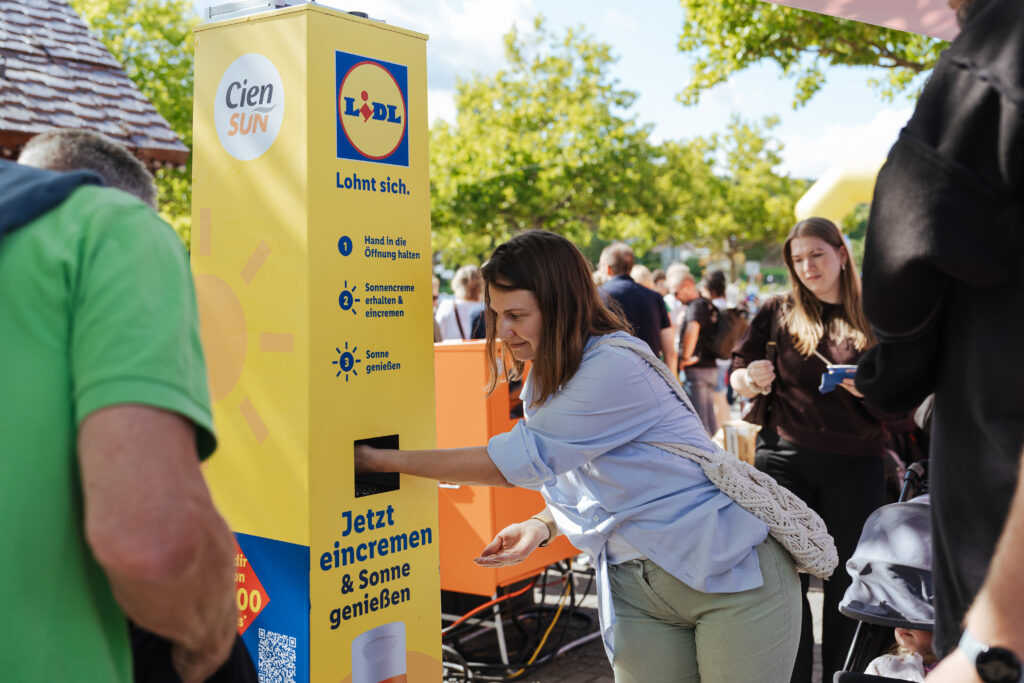 Frau hält ihre Hand in die Öffnung einer gelben Sonnencreme-Spendersäule von Lidl und Cien Sun und hält die andere Hand für die kostenlose Sonnencreme bereit, im Hintergrund weitere Besucher bei einer Veranstaltung im Freien