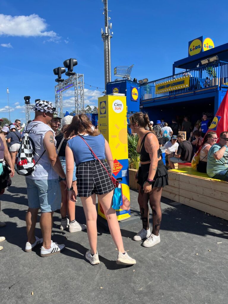 Young festivalgoers are lined up at a yellow sunscreen dispenser stand from Lidl and Cien Sun in front of the Lidl “Load Up Lounge” on a sunny festival grounds